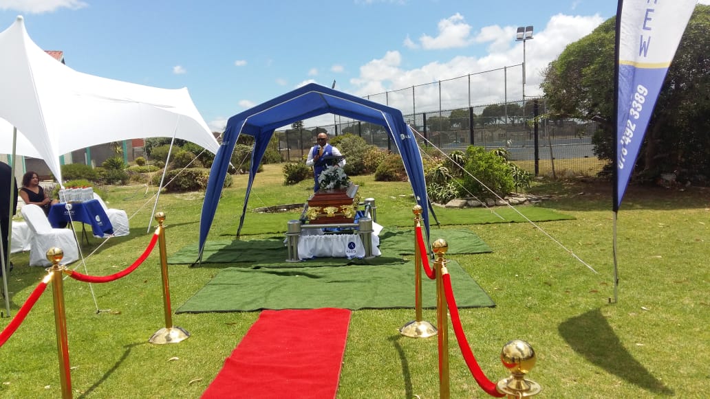 AMEW funeral service set up outdoors with a draped casket, floral tribute and family seating
